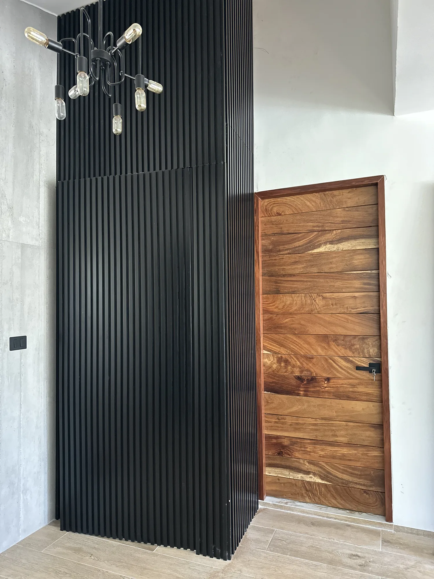 Lambrín de listones de madera en negro mate a doble altura junto a puerta de parota maciza